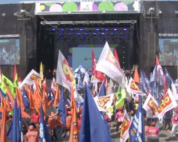 Bandeiras de centrais sindicais no ato de 1º de maio, Dia do Trabalho, realizado na Neo Química Arena, em São Paulo. Ao fundo, a marca da Petrobras, patrocinadora do evento